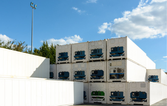 Refrigerated containers for Cedar Rapids IA stacked in a shipping yard. Trees and a blue sky with clouds peek out behind. 